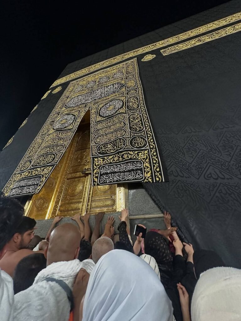 Umrah Travel Guide: Stress-Free & Reliable Transport A close-up low-angle view of pilgrims in Ihram touching the golden door and Kiswah of the Holy Kaaba at night.